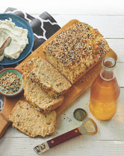Load image into Gallery viewer, Loaf of bread with slices on a wooden board, accompanied by a bottle of beer and condiments.