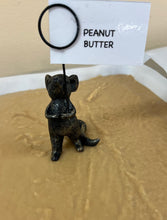 Load image into Gallery viewer, A small black metal figurine of a seated cat holding a sign that reads 'PEANUT BUTTER,' placed on a tan textured surface with a white tray edge in the background.