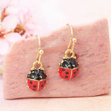 Load image into Gallery viewer, Red and black ladybug earrings with gold accents on a pink stone with blurred floral background