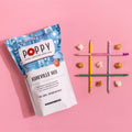 A bag of Poppy Hand-Crafted Asheville Mix popcorn sits on a pink background next to three colored pencils and scattered popcorn kernels.
