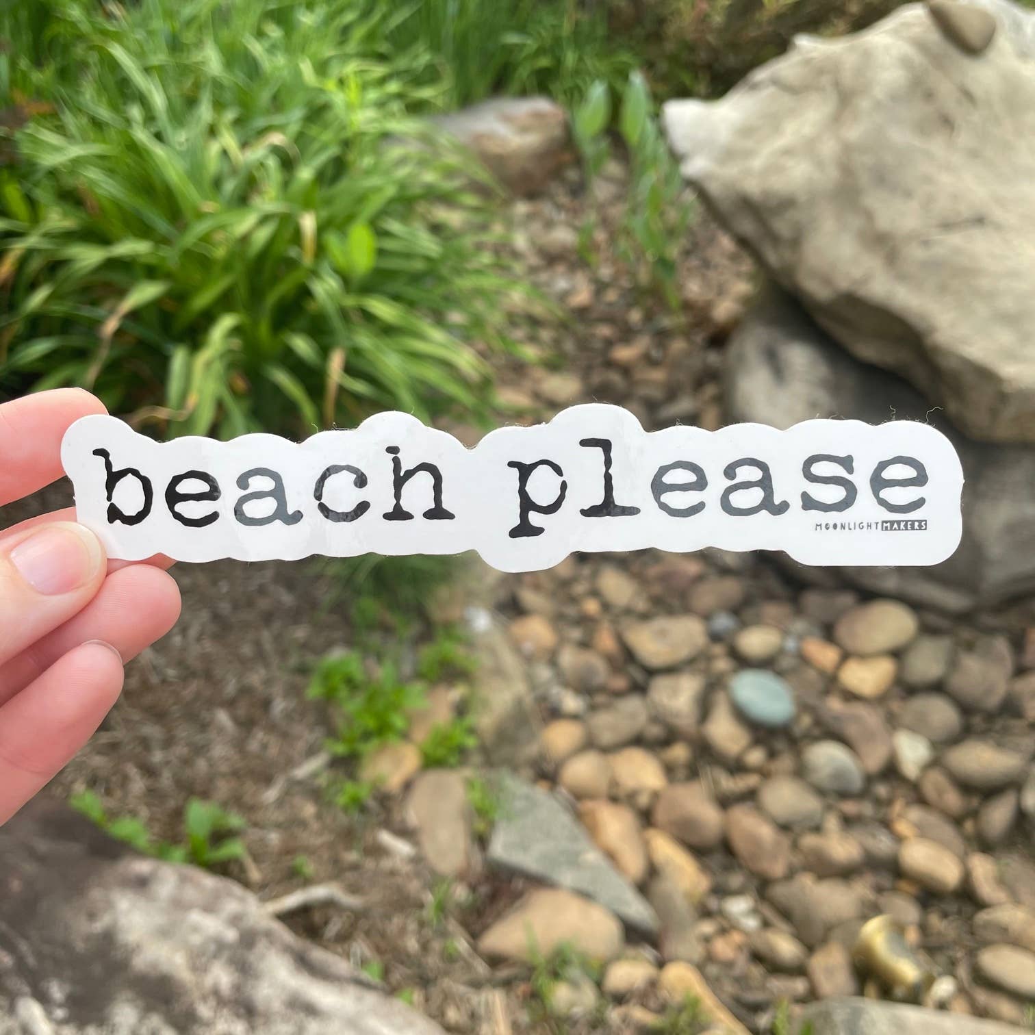 A hand holds a die-cut white sticker with the words 'beach please' in black lowercase-style lettering, with a small 'MORRISLY LEON' mark on the right side. The background is a blurred outdoor garden scene with rocks, soil, and green plants.