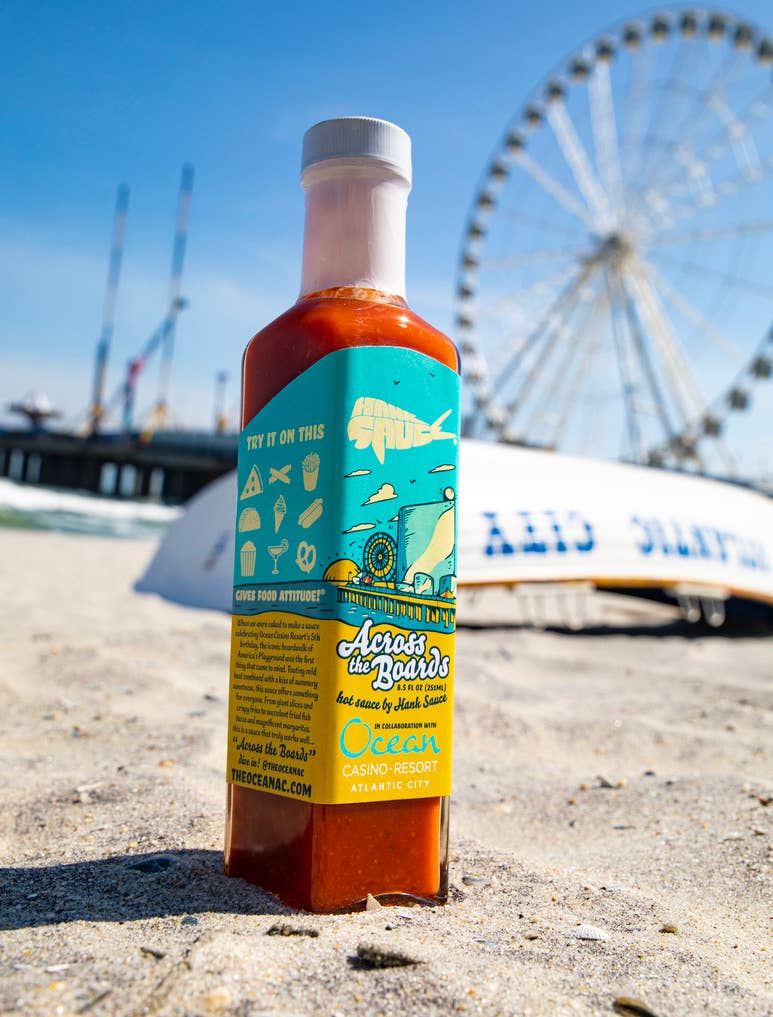 Bottle of Across the Boardwalk hot sauce on a beach with a Ferris wheel in the background
