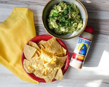 Load image into Gallery viewer, Bowl of guacamole with tortilla chips and a bottle of hot sauce on a wooden surface.
