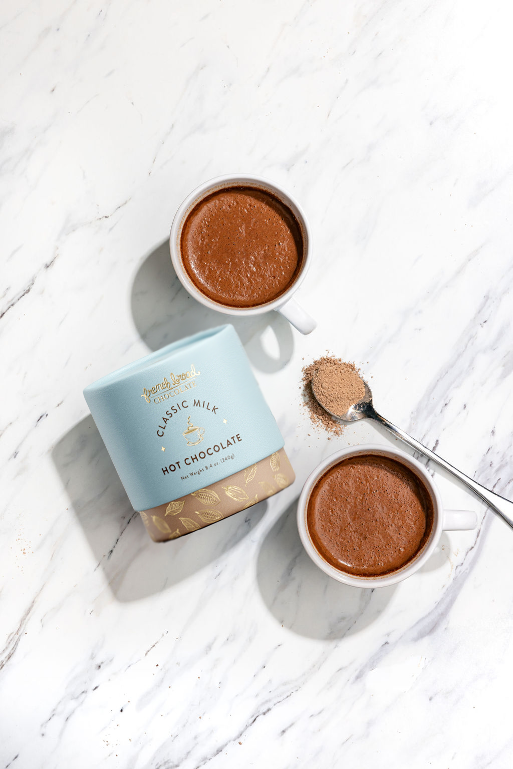 Two cups of hot chocolate with a box labeled 'Classic Milk Hot Chocolate' on a white wooden surface.
