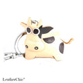 A small cow-shaped keychain charm made of light tan leather with black spots, large round cartoon-style eyes, and silver metal keyring hardware, shown on a white background. The word LeatherChic appears in the lower-left corner.