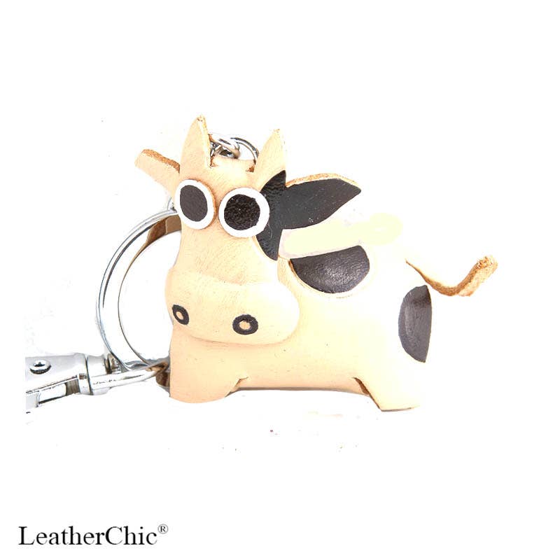 A small cow-shaped keychain charm made of light tan leather with black spots, large round cartoon-style eyes, and silver metal keyring hardware, shown on a white background. The word LeatherChic appears in the lower-left corner.