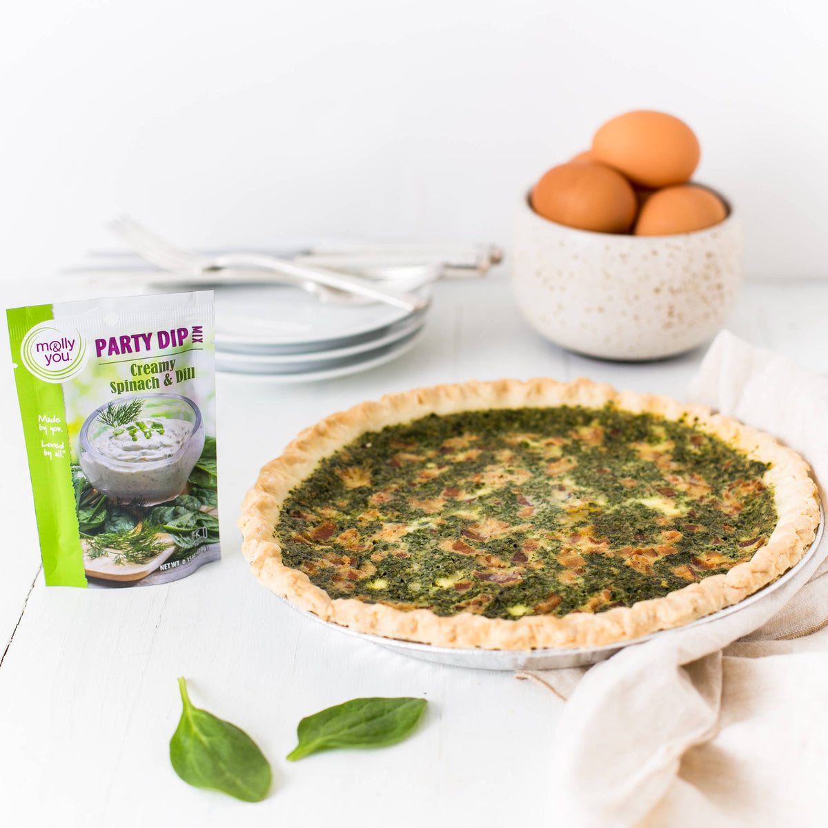 Spinach quiche on a white surface with a package of Party Dip and a bowl of eggs in the background.