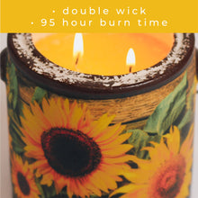 Load image into Gallery viewer, Candle with sunflower design and two wicks burning, on a yellow background with text about burn time.
