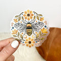 Decorative sticker with a bee and flowers held by a hand on a neutral background