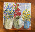 A square painted canvas or tile featuring three vases of flowers against a window-like background, placed on a wooden floor. The artwork shows blue flowers on the left, red flowers in the center, and yellow flowers on the right, with visible brushstrokes and a textured surface.