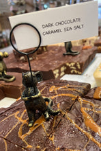 Load image into Gallery viewer, A close-up of a chocolate bar display with a small sign reading 'DARK CHOCOLATE CARAMEL SEA SALT.' The foreground piece is dark brown chocolate topped with caramel drizzle and coarse sea salt, with a small metallic figurine holding the sign.
