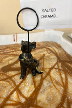 Load image into Gallery viewer, A small black metal mouse figurine stands on a caramel-topped dessert, holding a circular loop with a handwritten sign that reads 'SALTED CARAMEL'.
