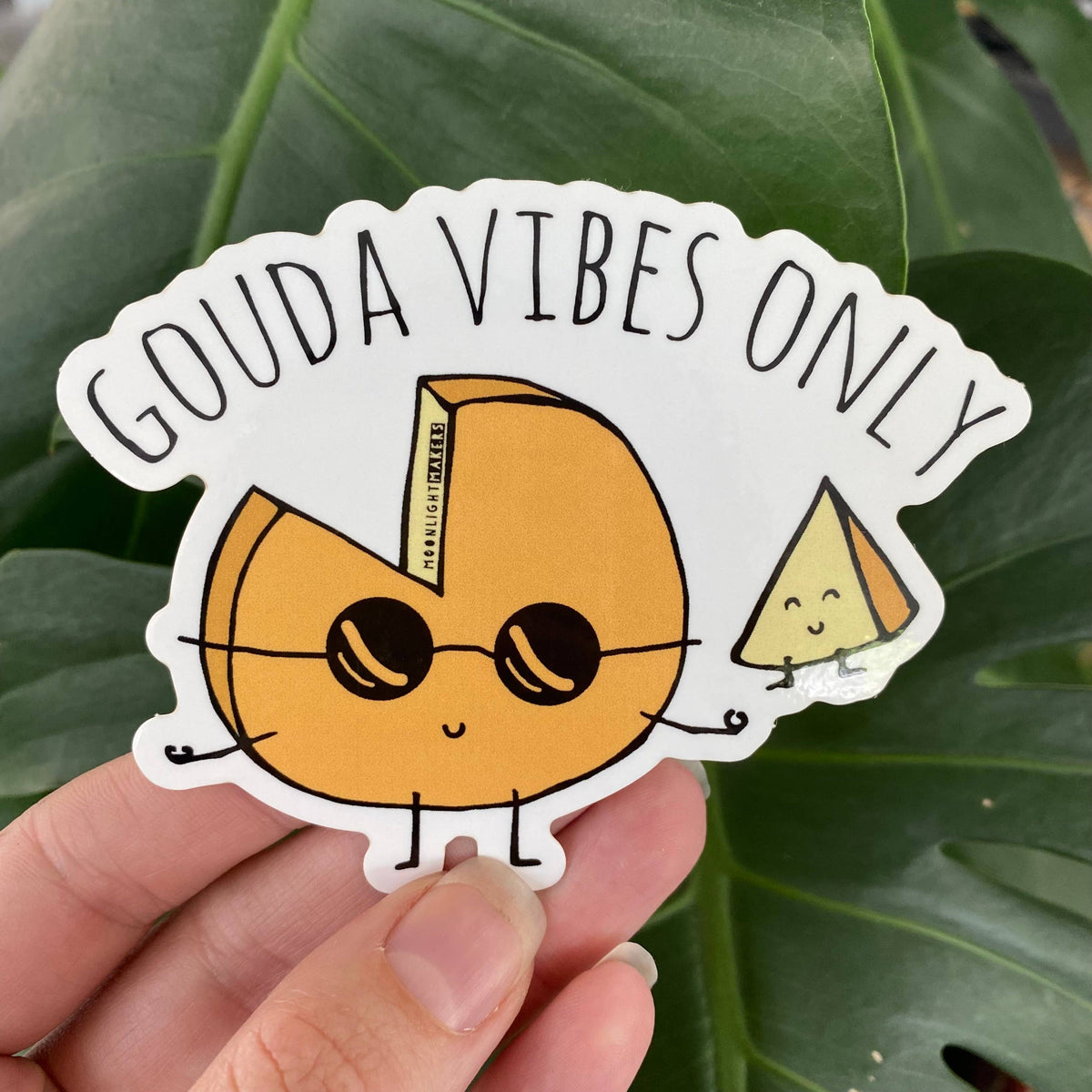 A hand holds a die-cut sticker featuring a cartoon wedge of cheese with sunglasses and the text 'GOUDA VIBES ONLY' against a background of large green leaves.