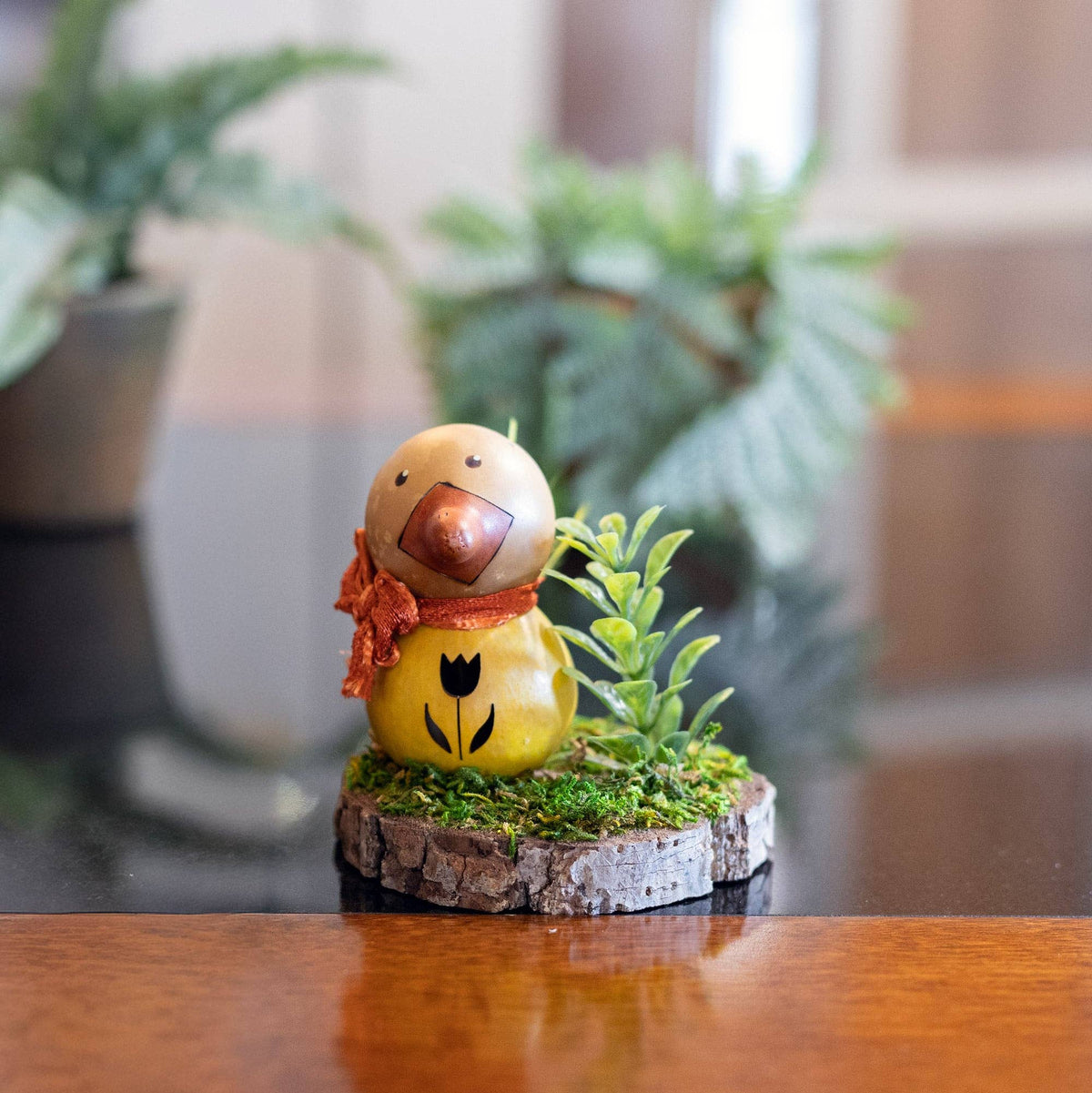 Decorative figurine of a duck with a red scarf on a wooden surface with plants in the background