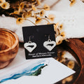Heart-shaped earrings on a 'House of Morgan Pewter' card with flowers and a wooden bowl in the background.