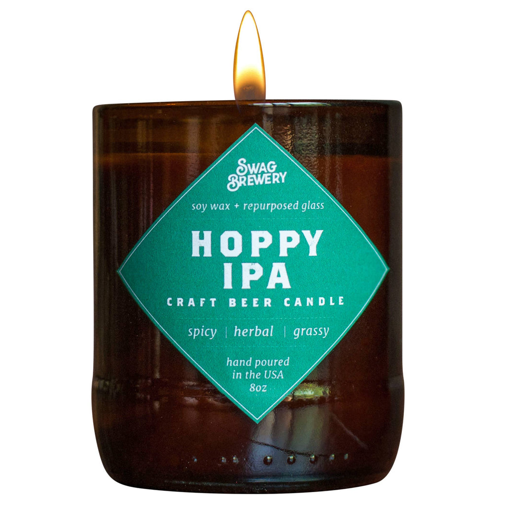 A lit Swag Brewery Hoppy IPA craft beer candle in a dark amber glass jar with a bright teal diamond-shaped label on the front.