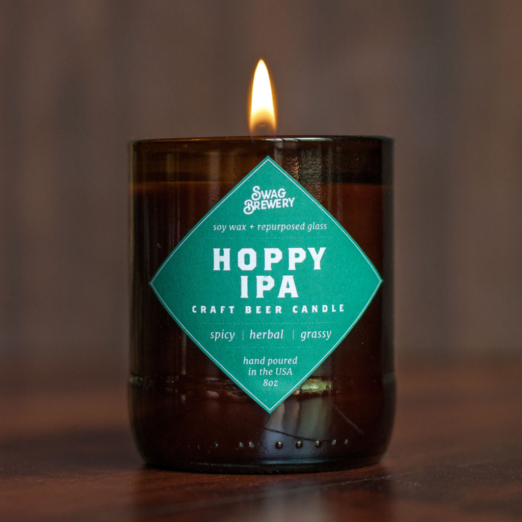 A lit Swag & Brewery Hoppy IPA craft beer candle in a dark amber glass jar, set on a wooden surface with a warm, blurred background.