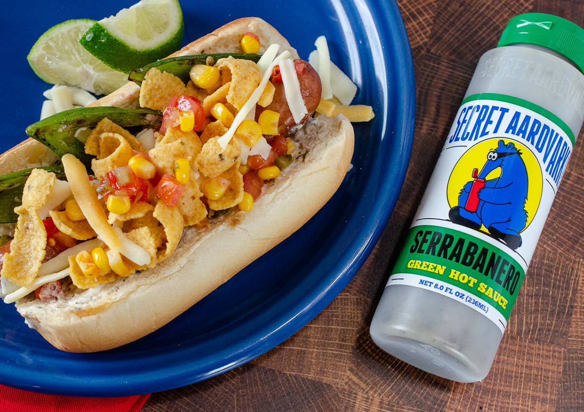 Hot dog with various toppings on a blue plate next to a bottle of Secret Aardvark Serrabanero green hot sauce.