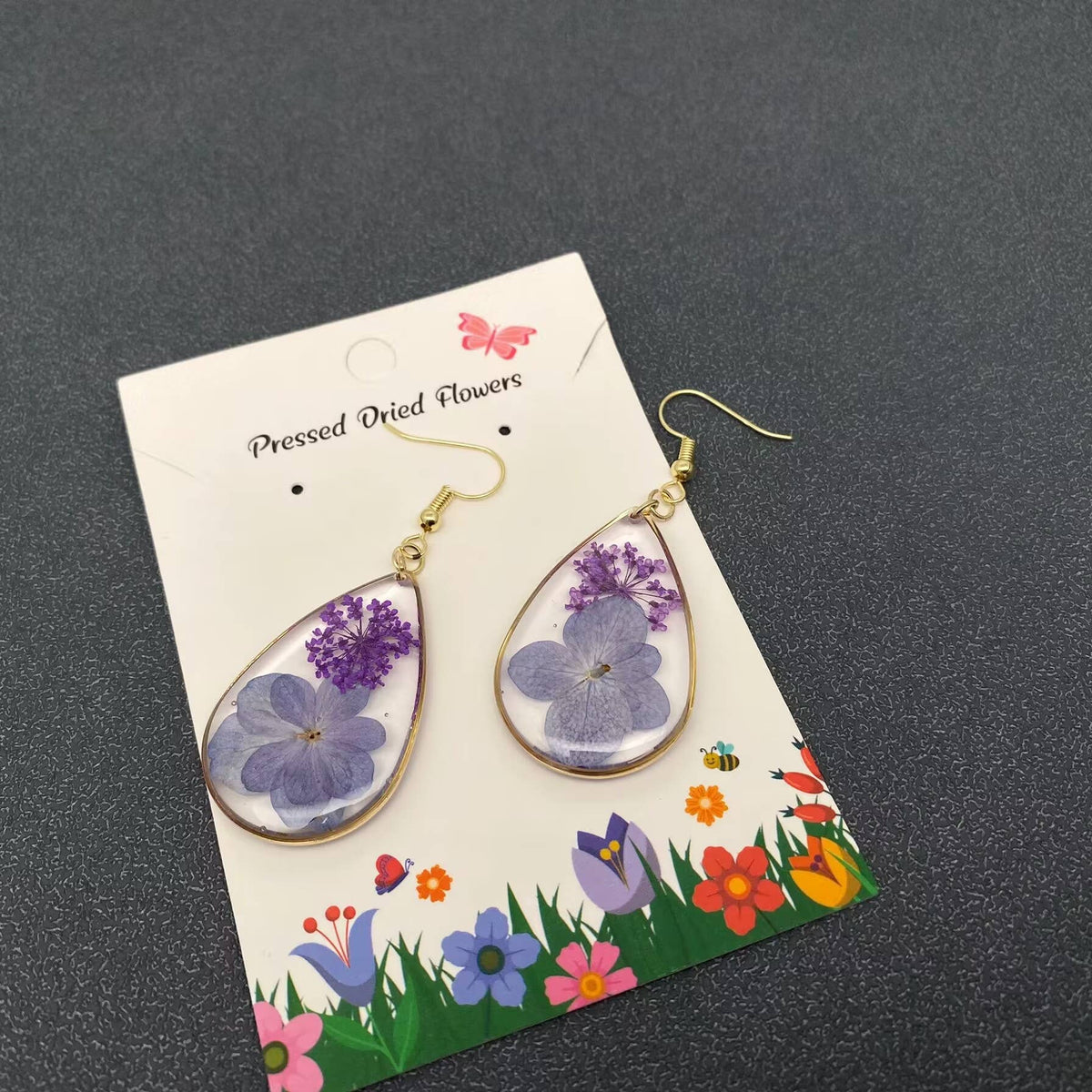 Teardrop-shaped earrings with pressed dried flowers on a decorative card against a dark background