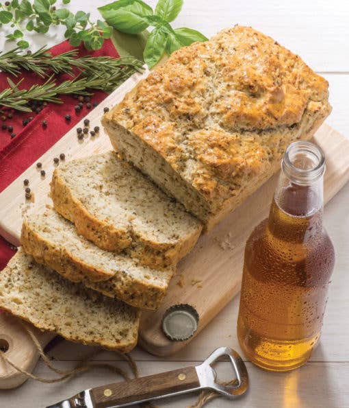 Loaf of bread sliced on a cutting board with a bottle of beer and herbs in the background.