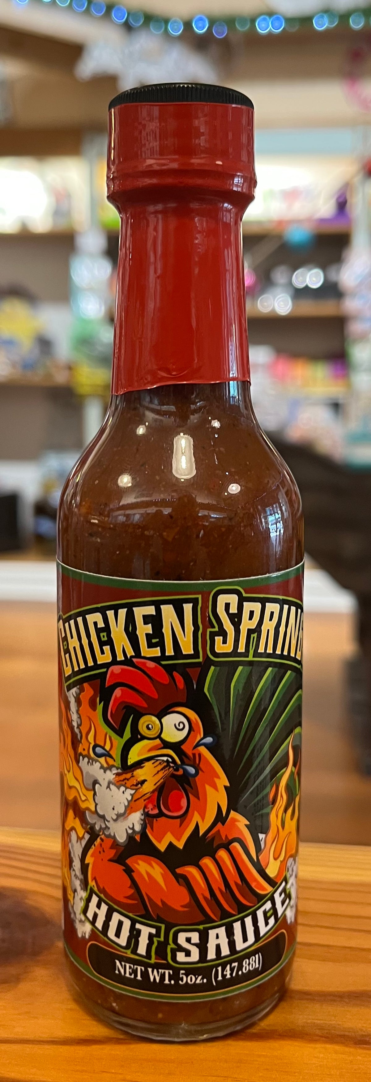 A bottle of Chicken Spring Hot Sauce on a wooden table with a blurred store background. The bottle has a red cap and a colorful label featuring a cartoon rooster.