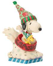 Load image into Gallery viewer, A decorative figurine of Snoopy wearing a green and red striped elf hat and a striped sweater, sitting in a red bowl on a wave-like base with a blue striped bottom. A small yellow bird is perched on Snoopy's shoulder.
