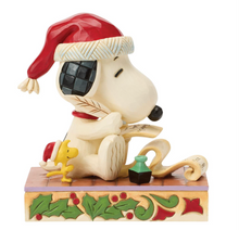 Load image into Gallery viewer, A decorative figurine of Snoopy wearing a red Santa hat and reading a scroll, with Woodstock beside him and a holly-accented base, photographed on a plain white background.
