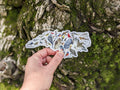 Hand holding a nature-themed sticker with various animals against a tree bark background