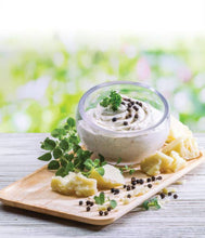 Load image into Gallery viewer, Bowl of cream cheese spread with herbs and black pepper on a wooden board with a blurred green background
