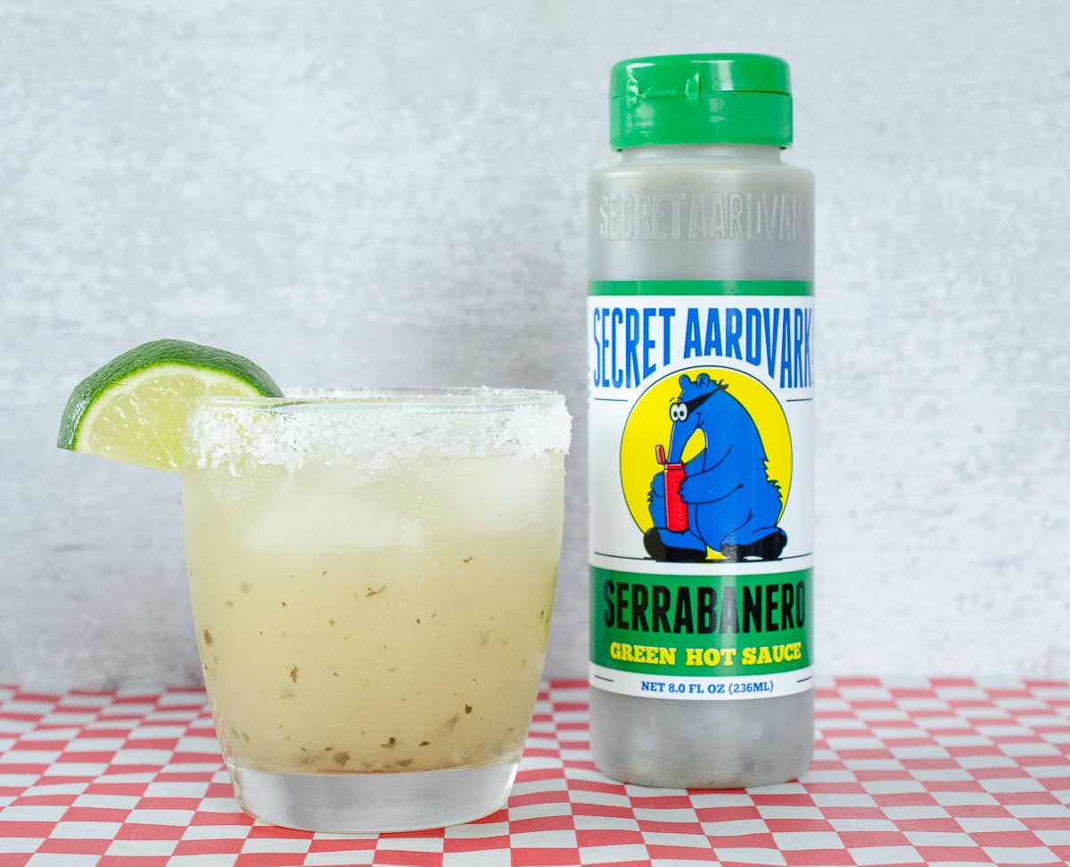 Bottle of Secret Aardvark Serranoblanco Green Hot Sauce next to a glass with a lime wedge on a checkered tablecloth.

