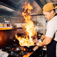 Load image into Gallery viewer, Person cooking with a large flame in a kitchen setting
