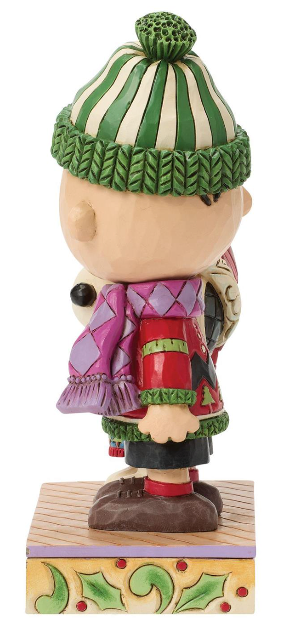 A collectible figurine of Charlie Brown from Peanuts, shown from the back wearing a green and cream striped knit hat, a purple argyle sweater, a red vest, and dark pants, standing on a decorated wooden base.