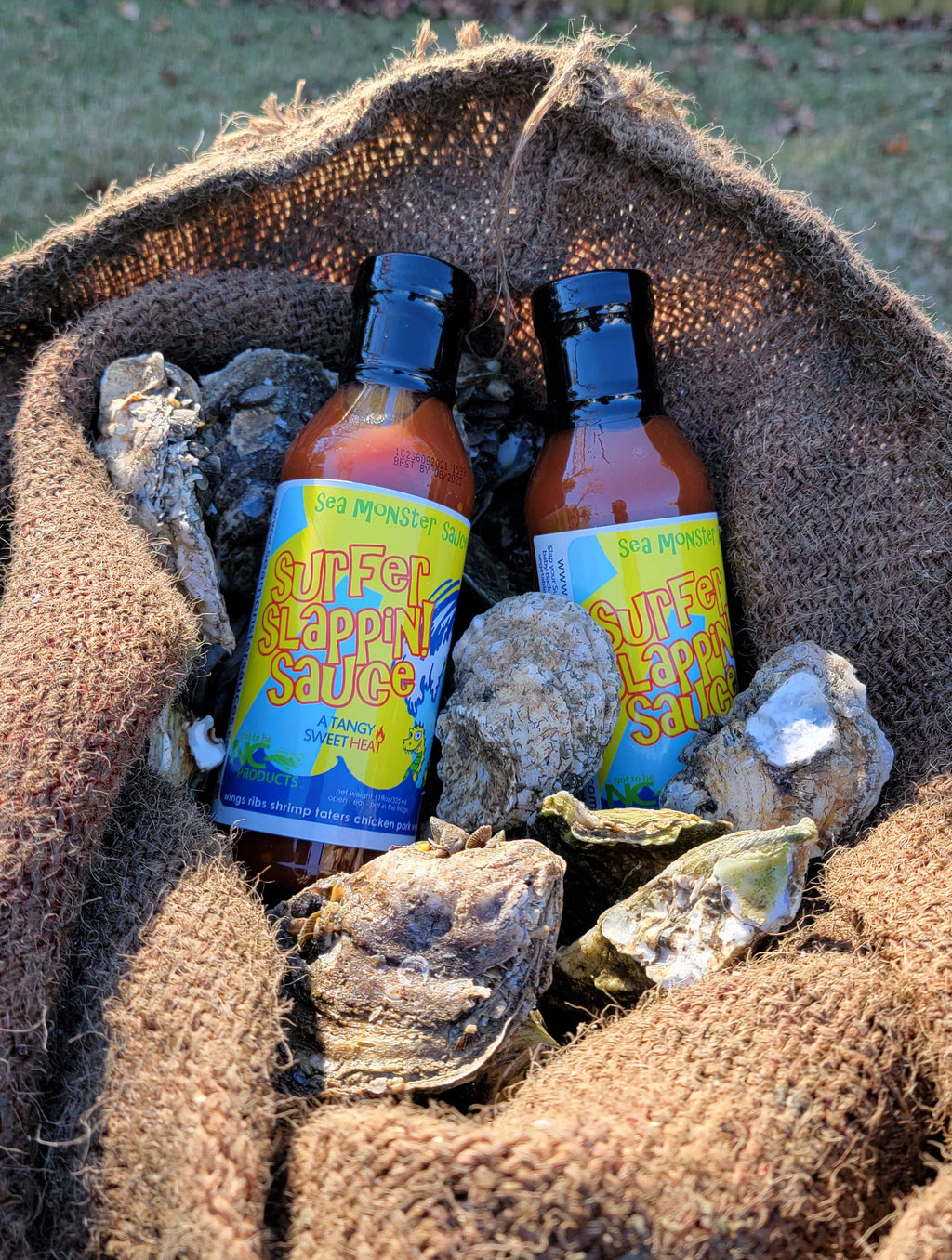 Two bottles of Surfer Slapping Sauce on a bed of oysters with a burlap bag in the background.