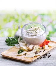 Load image into Gallery viewer, Dip in a glass bowl with garlic, herbs, and peppers on a wooden board with a blurred green background.
