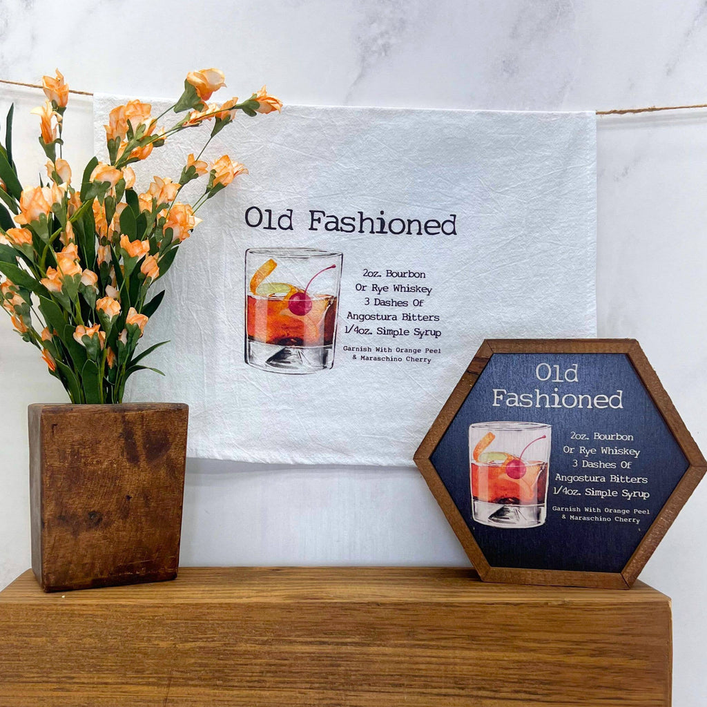 Decorative signs with 'Old Fashioned' cocktail recipe and illustration on a wooden surface with flowers.