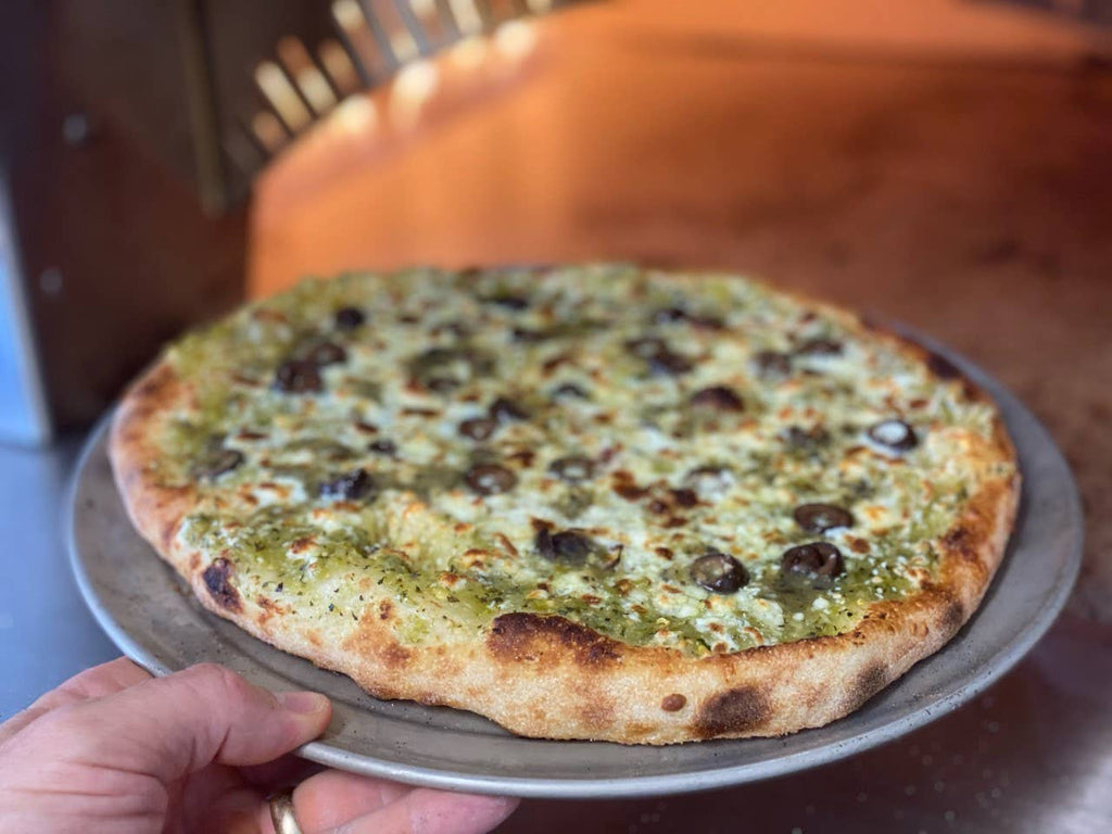 Person holding a pizza with olives and green sauce on a metal tray.